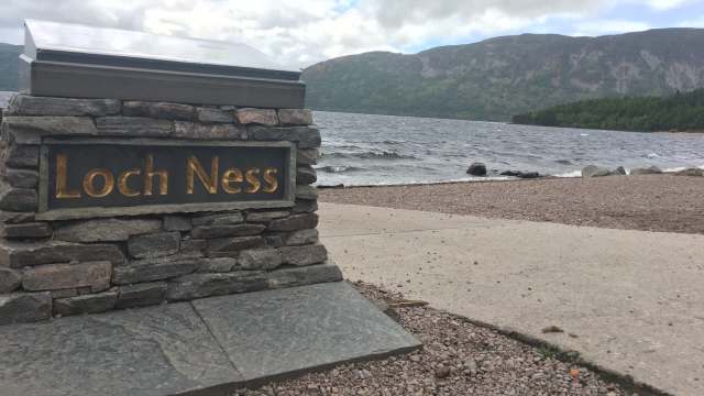 A stone sign reading "Loch Ness" by the lakeshore with waves and hills in the background.