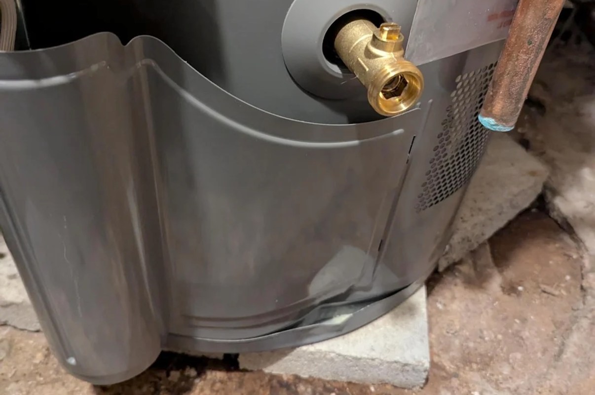 A close-up of a gray water heater, with a brass fitting on a concrete block in a basement, that has a dent at the bottom.