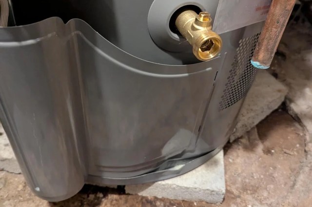 A close-up of a gray water heater, with a brass fitting on a concrete block in a basement, that has a dent at the bottom.