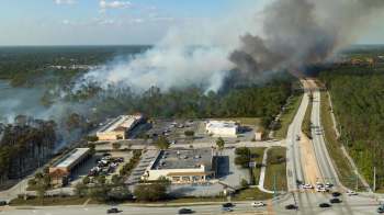 Aerial view of smoke rising from a wooded area near a commercial district and road.