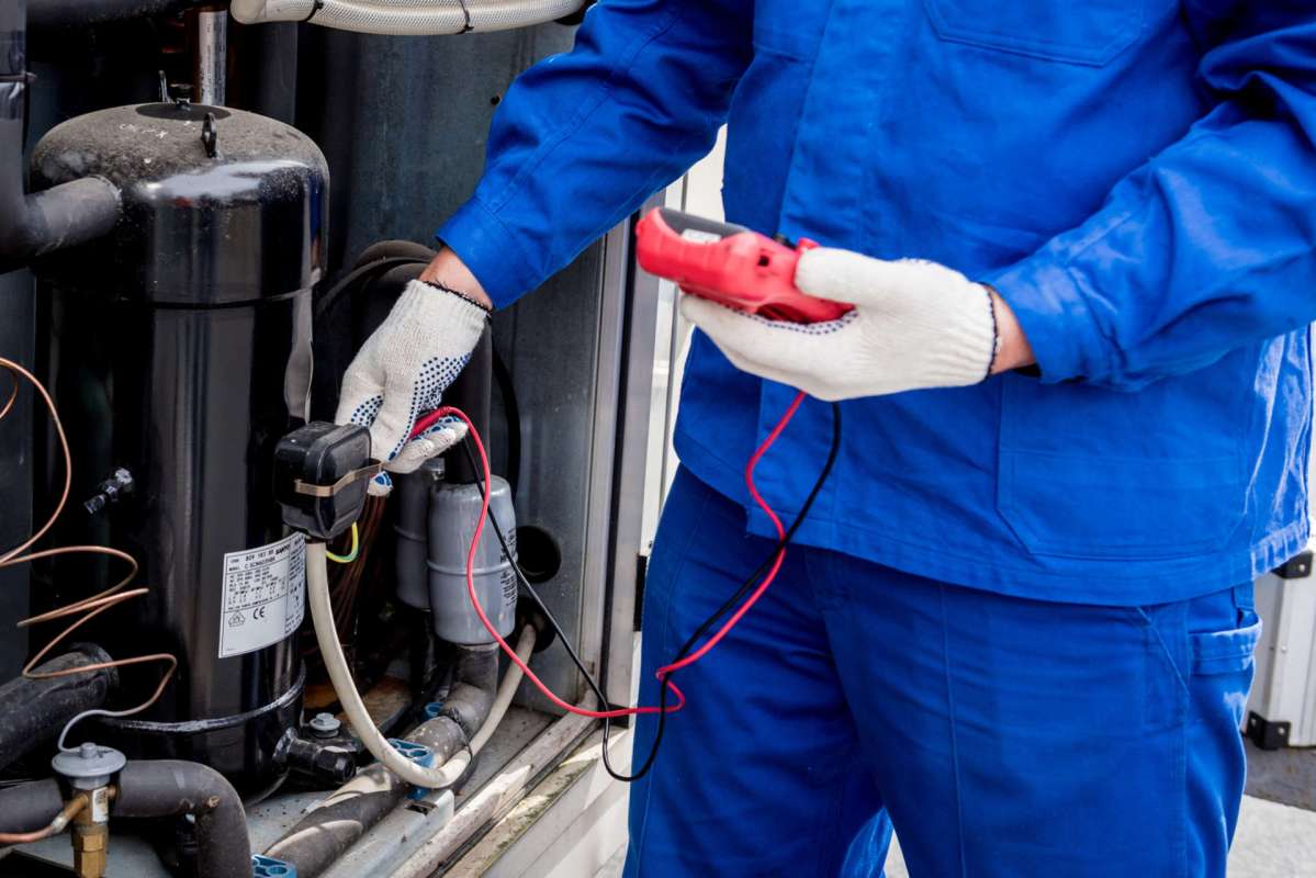 A technician tests a compressor with a multimeter while wearing gloves.