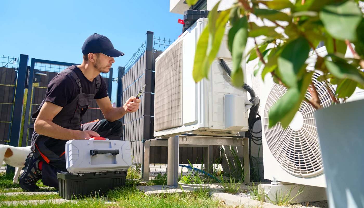 A technician examines an outdoor air conditioning unit while a small dog sits nearby.