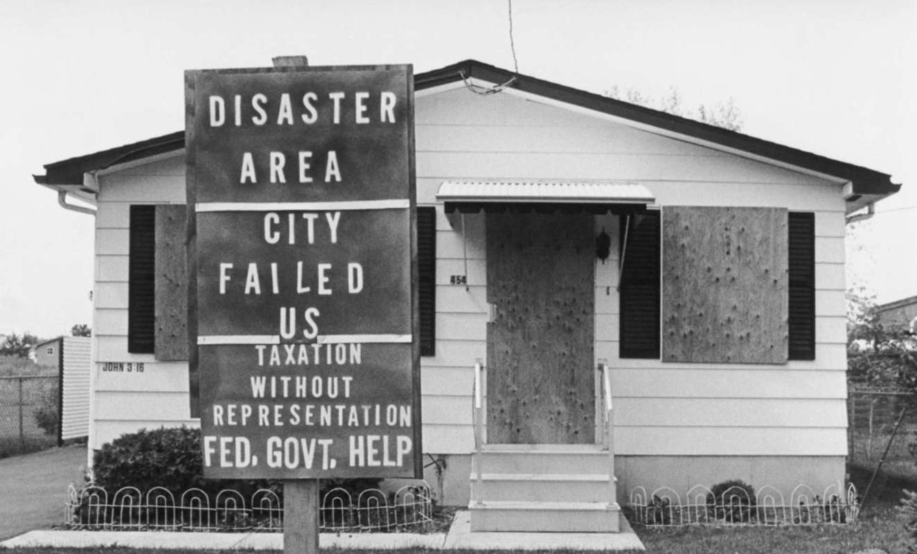 A house with a boarded-up entrance and a sign declaring it a disaster area seeking government help.