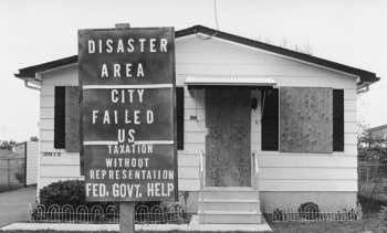 A house with a boarded-up entrance and a sign declaring it a disaster area seeking government help.