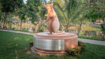 A wooden statue of a rat stands on a circular base in a park surrounded by greenery.