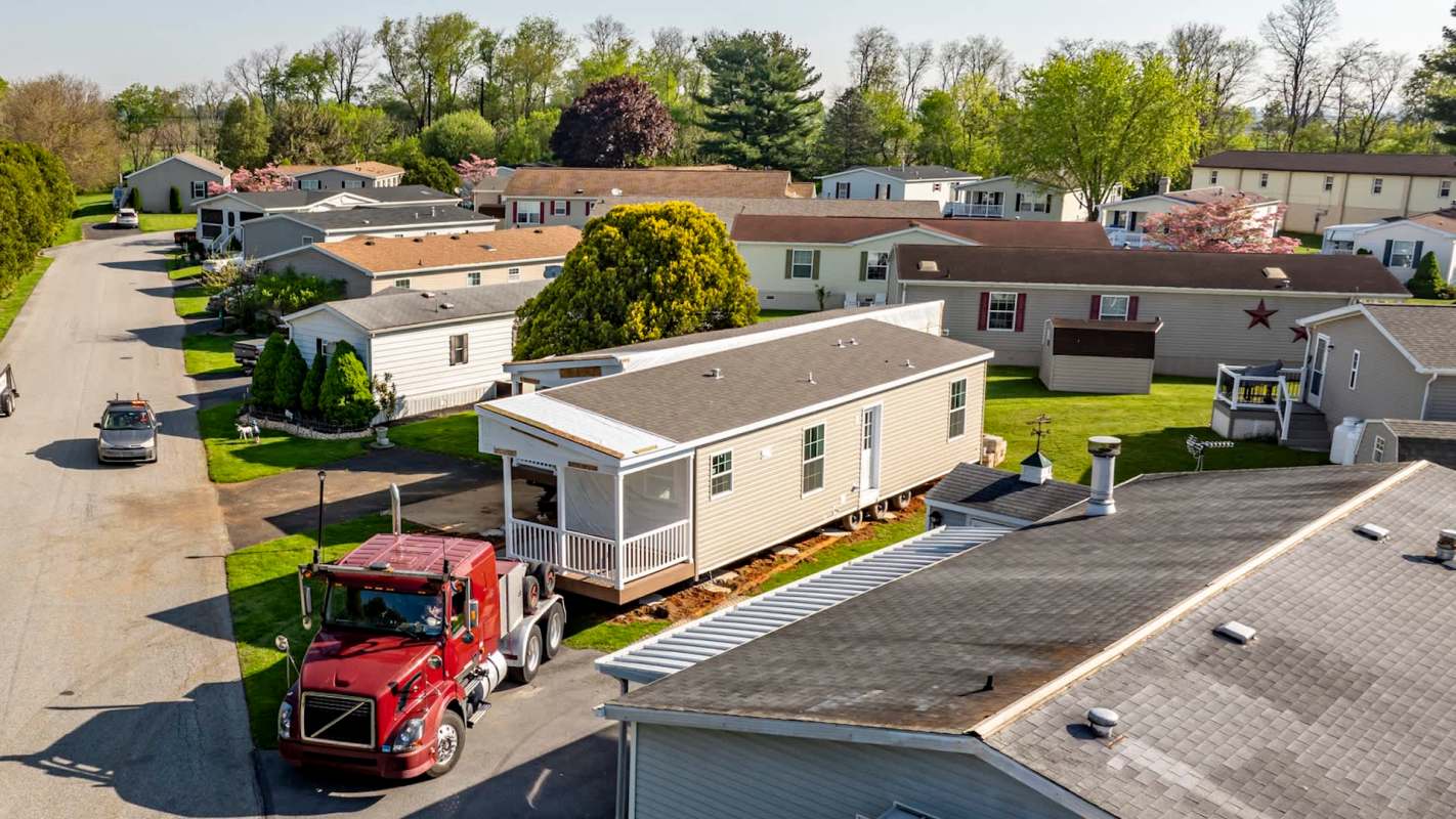 A red truck delivers a mobile home to a residential area with other homes and trees.