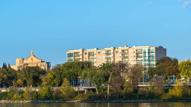 A stone building and a modern apartment complex surrounded by trees along a river under a clear blue sky.