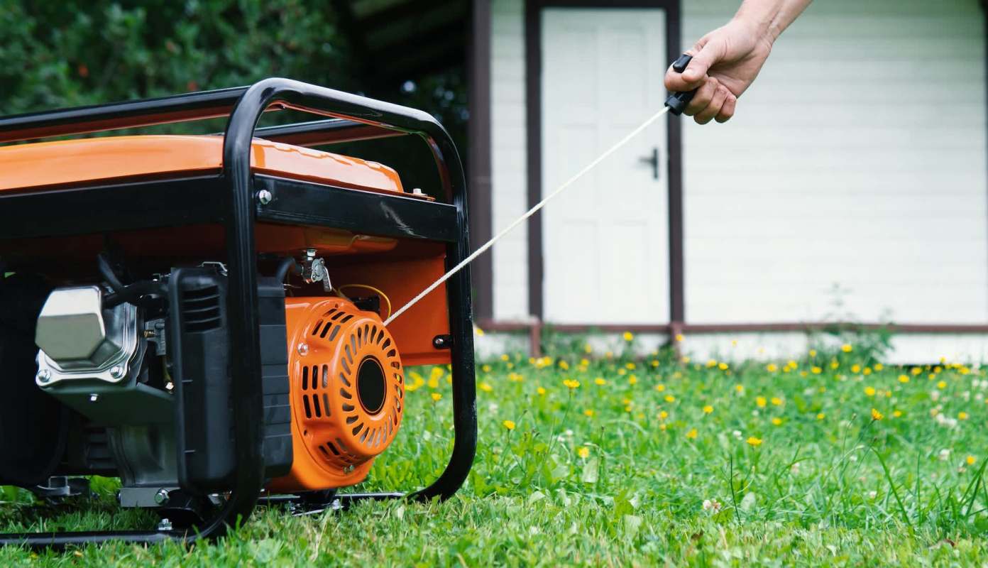 A person pulls the starter cord of a generator on a lawn near a house.