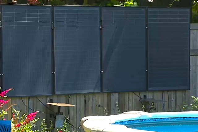 Solar panels installed beside a swimming pool, surrounded by plants and a wooden fence.