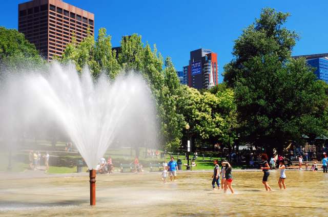 A fountain sprays water in a park where people walk and play in shallow water.