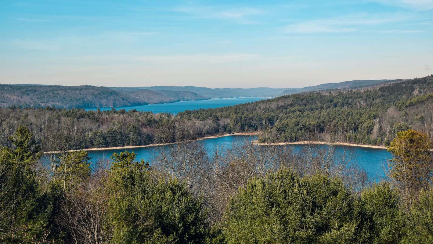 A panoramic view of a lake surrounded by forests and rolling hills under a clear blue sky.