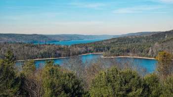A panoramic view of a lake surrounded by forests and rolling hills under a clear blue sky.