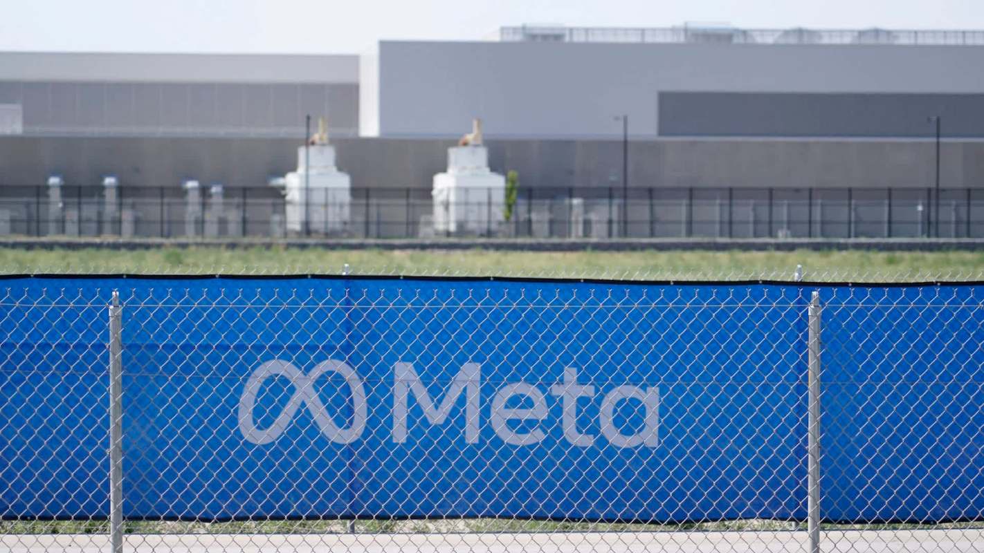 A blue fence with the Meta logo and a background of industrial buildings and green fields.