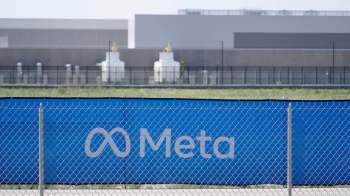 A blue fence with the Meta logo and a background of industrial buildings and green fields.