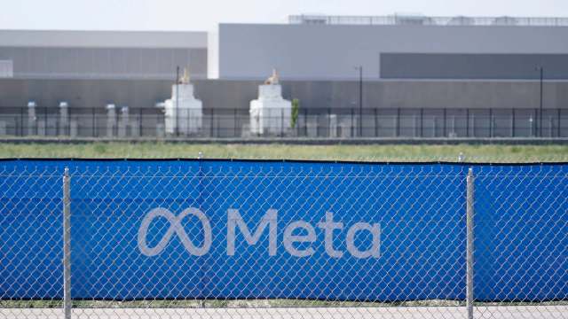 A blue fence with the Meta logo and a background of industrial buildings and green fields.