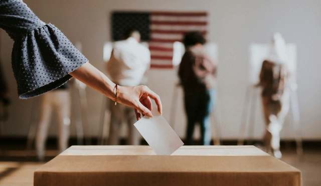 A hand places a ballot into a voting box with people voting in the background.