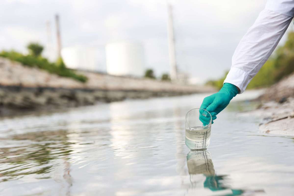 A person in gloves collects water samples from a shallow body of water near industrial structures.