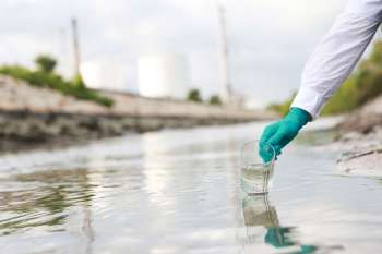 A person in gloves collects water samples from a shallow body of water near industrial structures.
