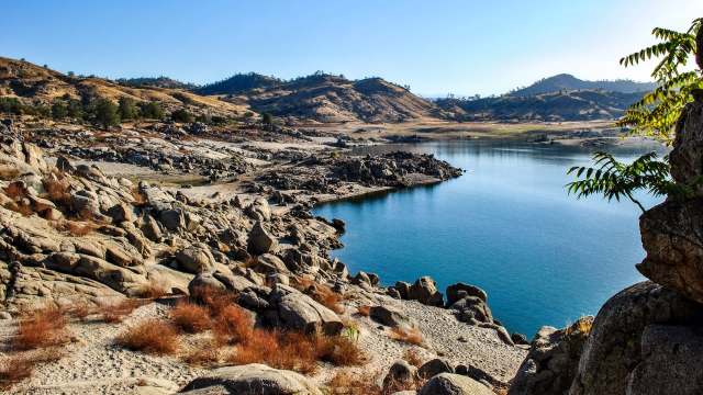 A rocky shoreline leads to a calm body of water surrounded by hills and sparse vegetation.