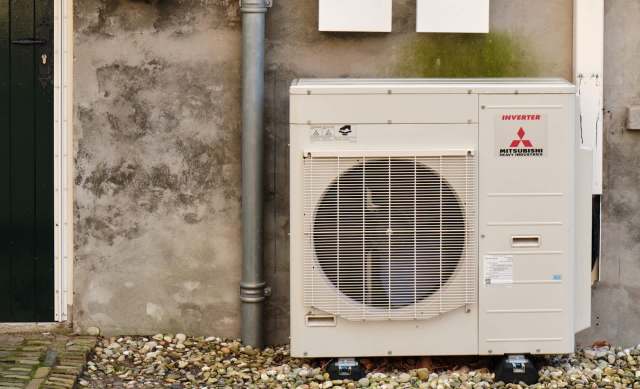 A Mitsubishi outdoor heat pump unit next to a wall, with a pipe and pebbles on the ground.