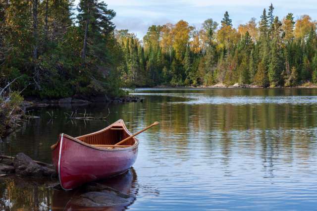 A red canoe next to a calm lake surrounded by trees with autumn foliage.
