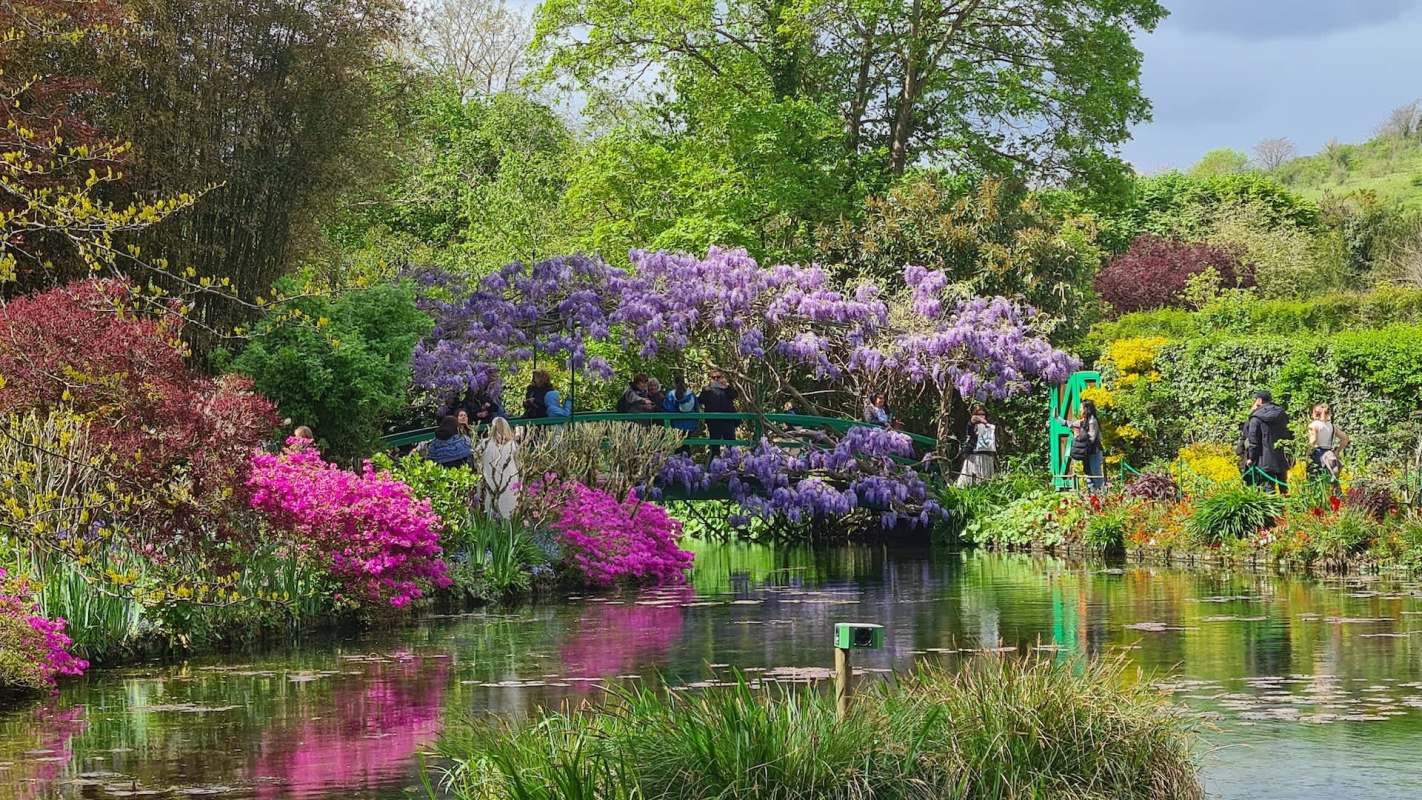 A garden with colorful flowers, a pond, and visitors on a bridge surrounded by lush greenery.