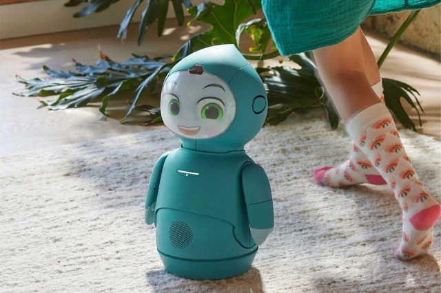 A teal robot with a smiling face stands on a carpet near a child’s foot wearing colorful socks.