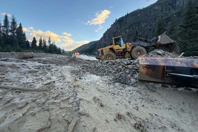 A yellow construction vehicle moves debris on a muddy road surrounded by trees and mountains.