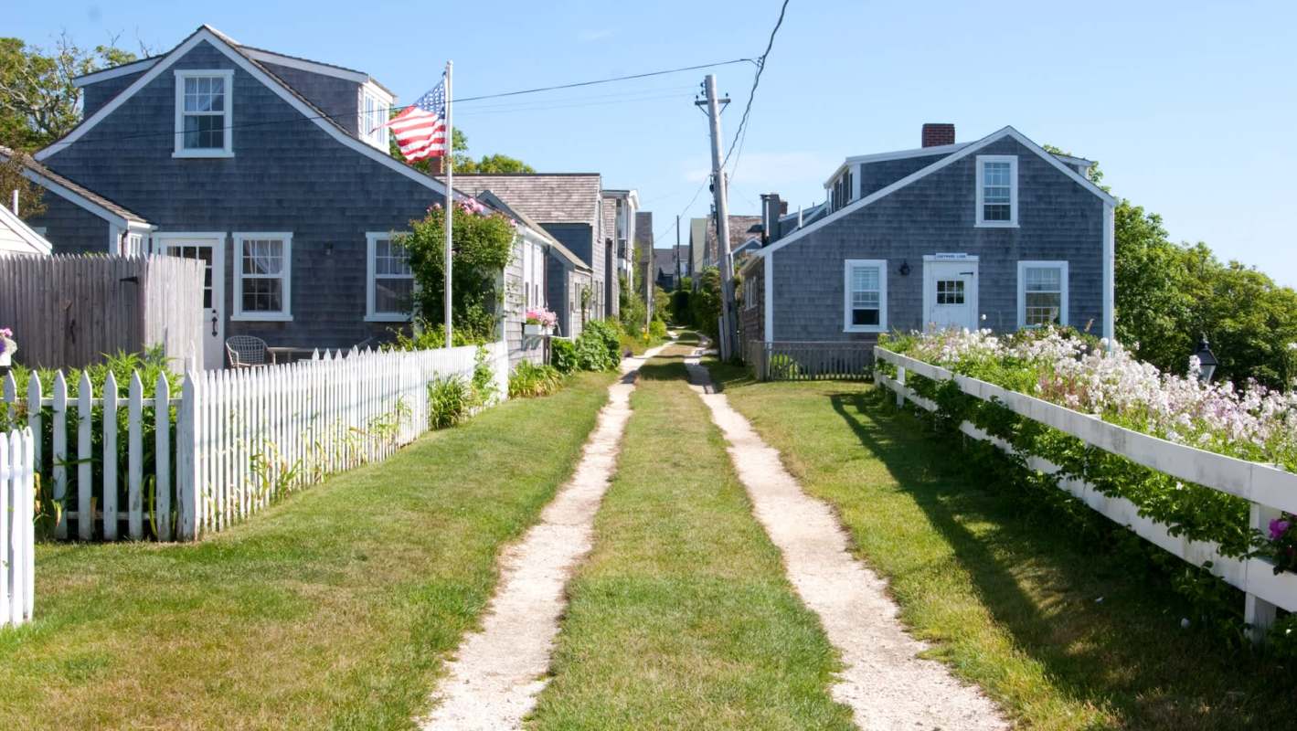 A dirt path between two houses with a white picket fence and flowers along the edges.