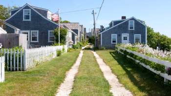 A dirt path between two houses with a white picket fence and flowers along the edges.