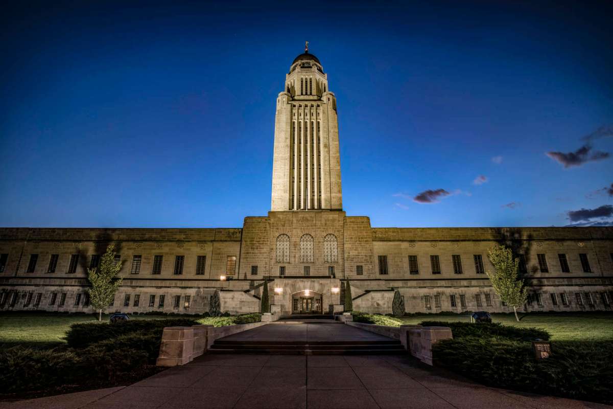 The Nebraska State Capitol building illuminated at dusk, with clear skies and landscaped grounds.