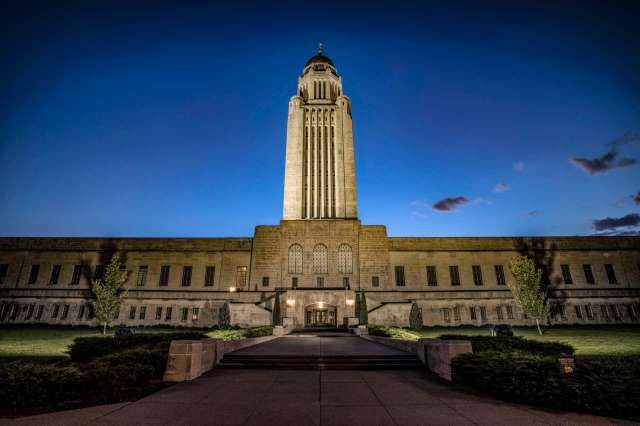 The Nebraska State Capitol building illuminated at dusk, with clear skies and landscaped grounds.