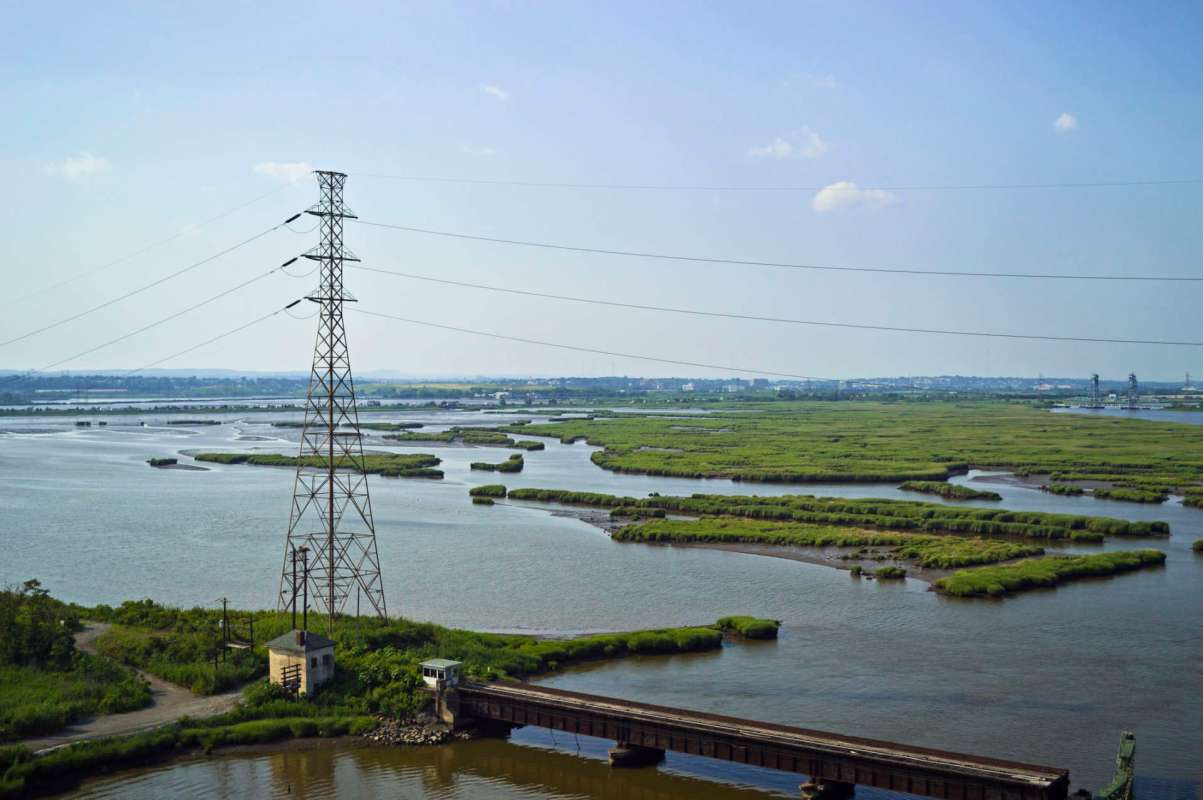 A power line towers over a winding river surrounded by green marshland and distant buildings.