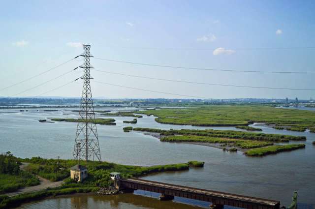 A power line towers over a winding river surrounded by green marshland and distant buildings.