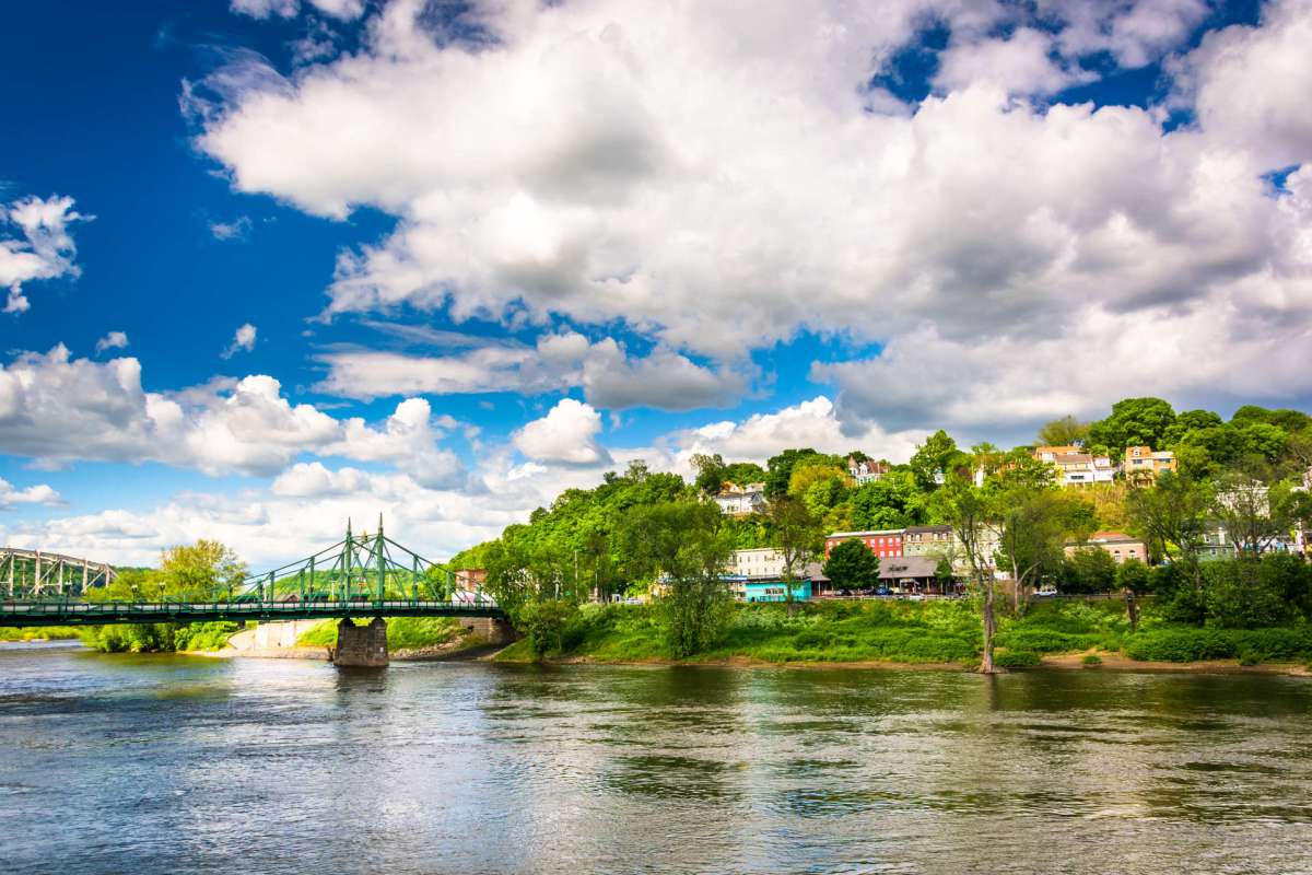 A green bridge crosses a river with a hillside of houses and trees under a blue sky filled with clouds.