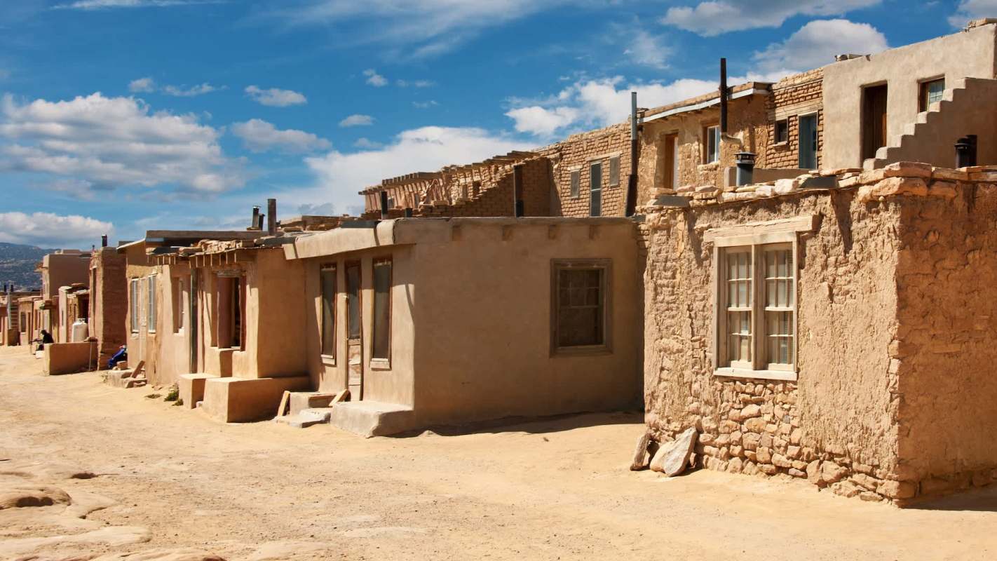 A dirt path lined with adobe buildings under a partly cloudy sky.