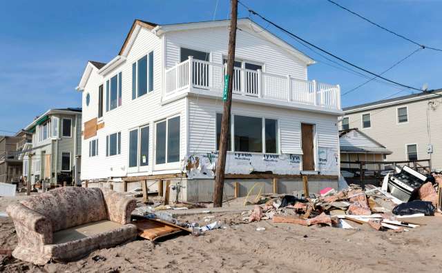 A damaged house sits among debris and sand, with a floral couch nearby and other broken items scattered around.