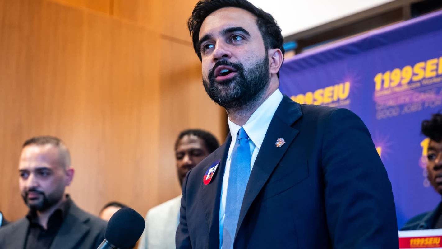 New York mayor Zohran Mamdani speaks at a podium with a union banner in the background, accompanied by several people.
