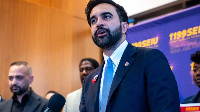 New York mayor Zohran Mamdani speaks at a podium with a union banner in the background, accompanied by several people.