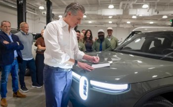 California Gov. Gavin Newsom signs a document on the hood of a Rivian vehicle, surrounded by several people in a workshop.