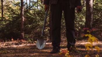 A person holding a shovel stands in a forest with trees and scattered foliage.