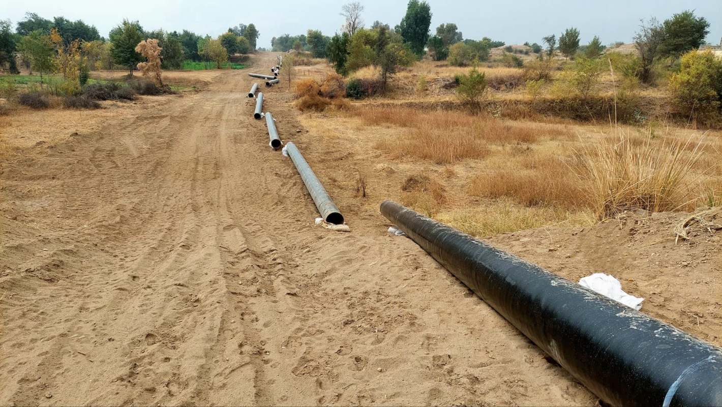 A series of long black pipelines laid along a dirt path in a dry, grassy area with sparse trees.