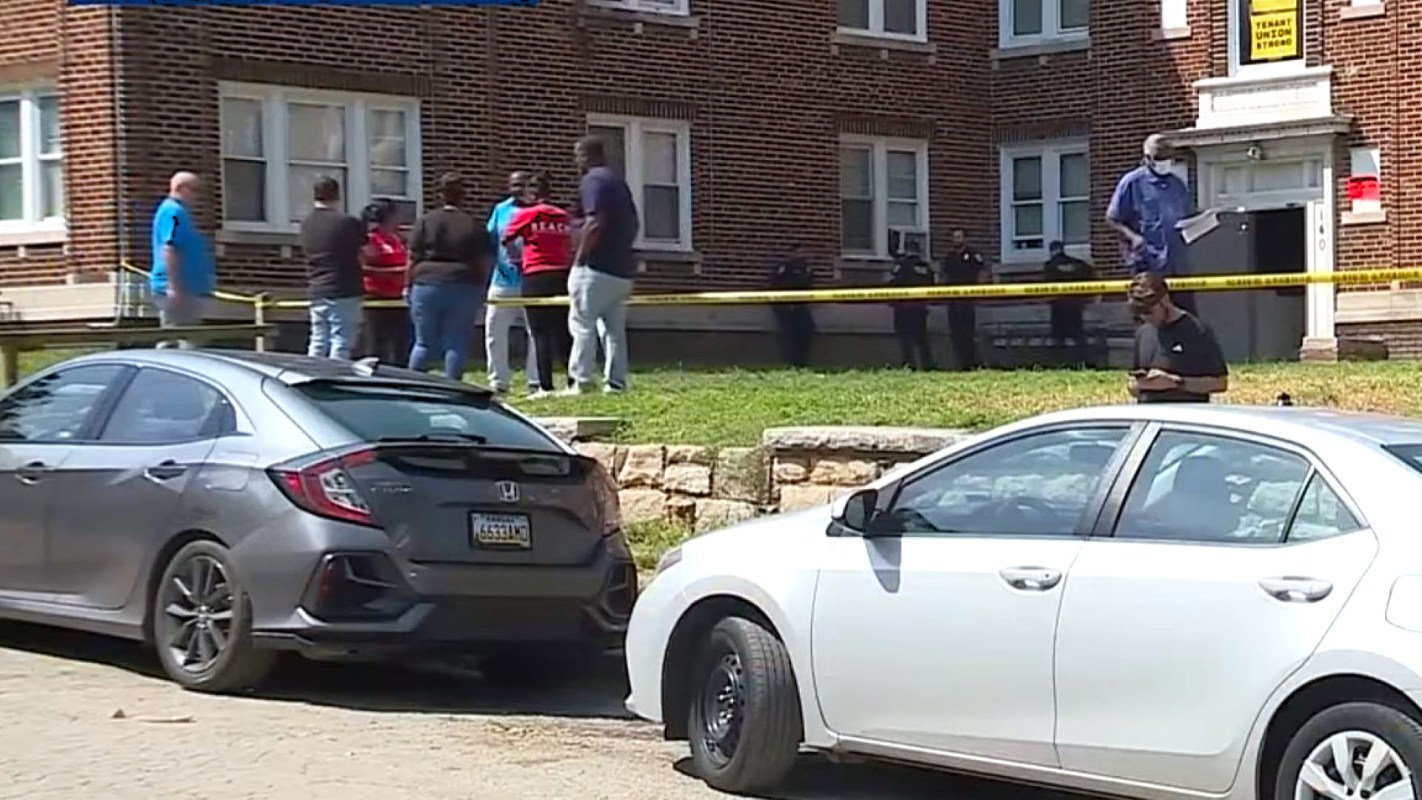 A group of people stands near an apartment building cordoned off by police tape.