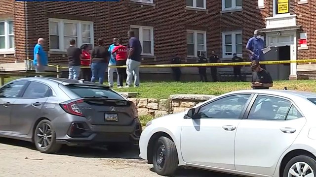 A group of people stands near an apartment building cordoned off by police tape.