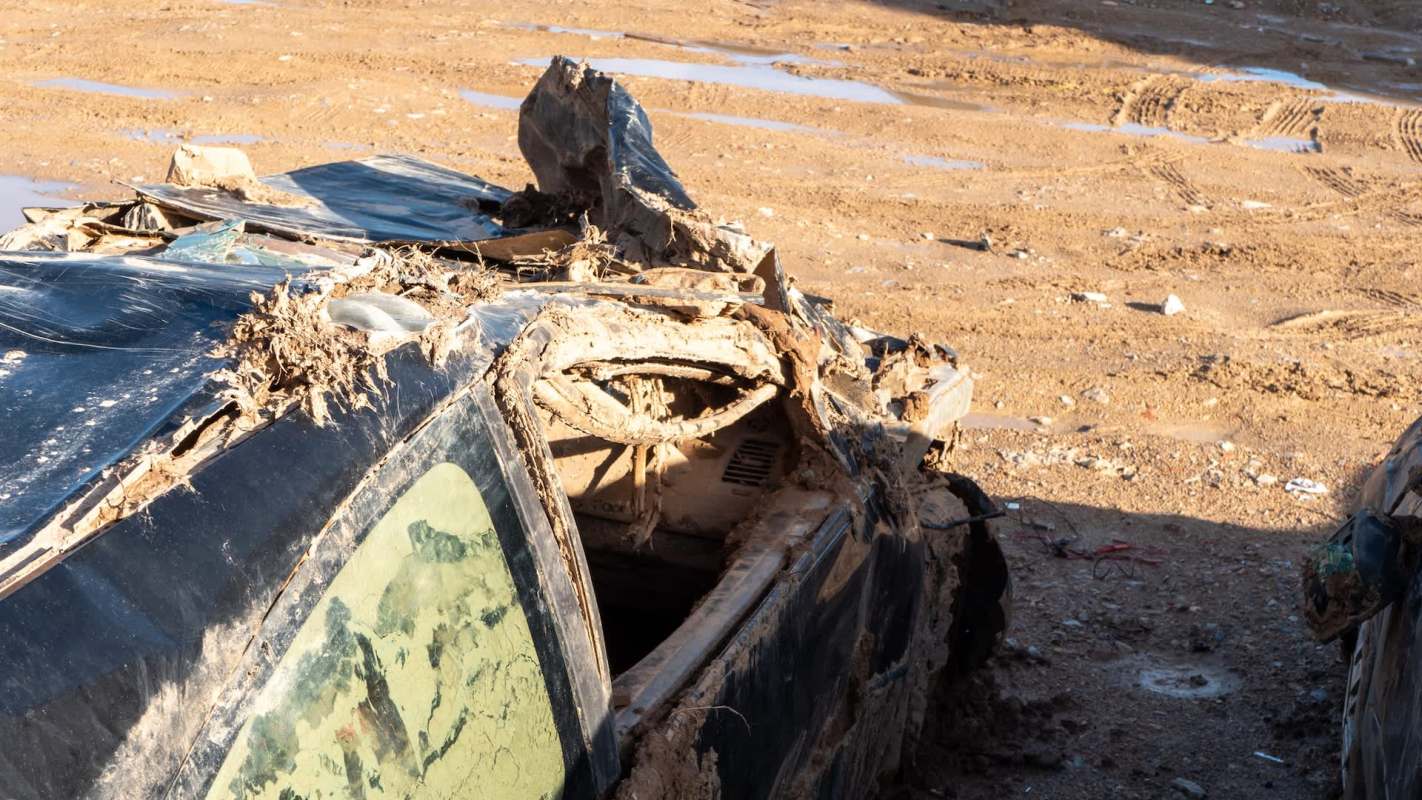 An abandoned, damaged car covered in dirt and debris in a muddy area.