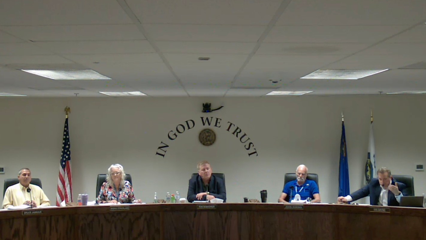 A group of five individuals is seated at a table with a wall displaying "In God We Trust."