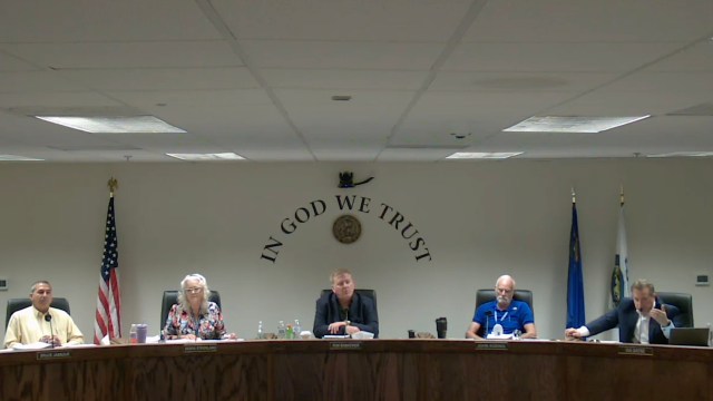 A group of five individuals is seated at a table with a wall displaying "In God We Trust."