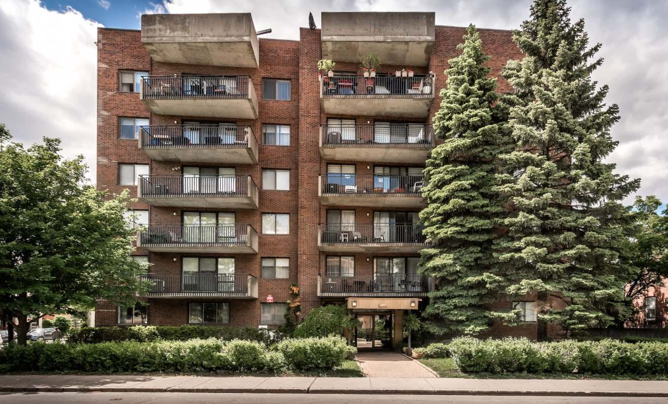 A multistory brick apartment building with balconies and surrounding greenery.