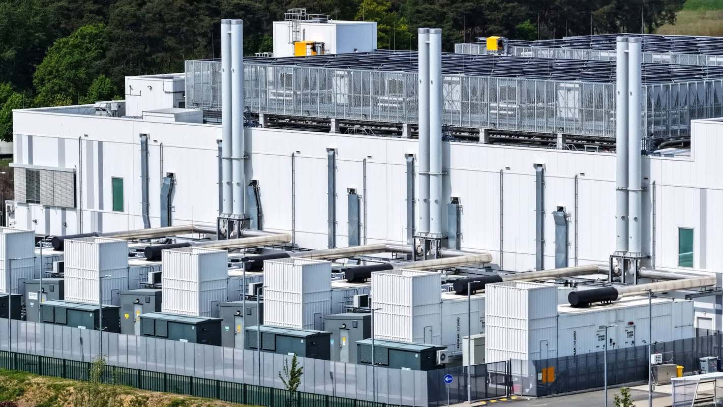 A building with large cooling units and exhaust pipes surrounded by a fence and green trees.