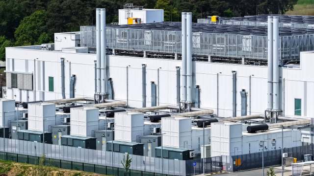 A building with large cooling units and exhaust pipes surrounded by a fence and green trees.